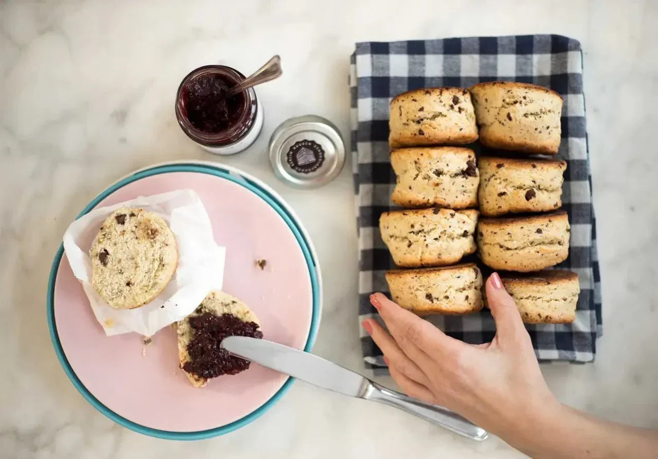 Frisch gebackene Schoko-Scones mit Marmelade auf einem rosa Teller und einer blauen Serviette auf Marmoruntergrund.