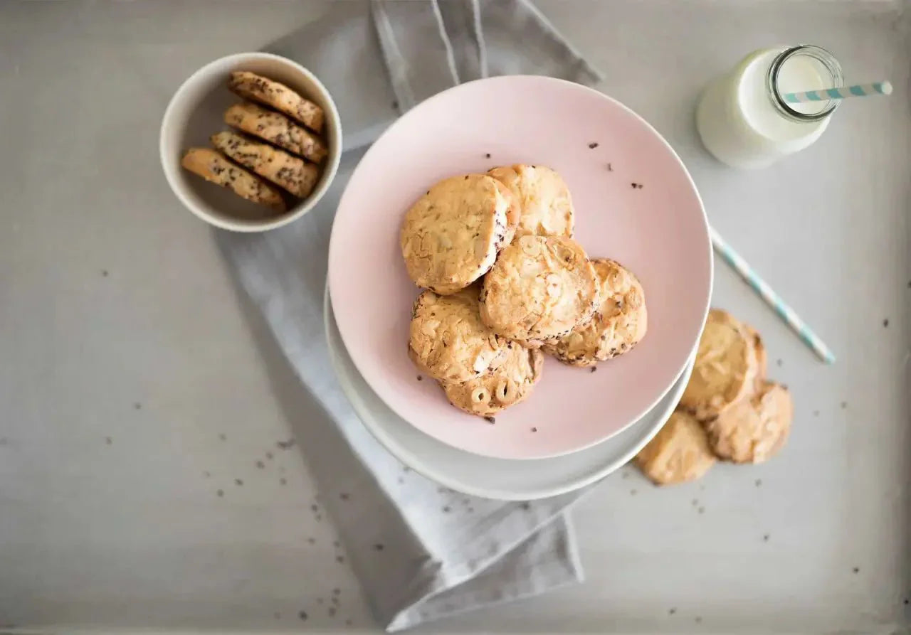 Cookies mit weißer Schokolade, Nüssen und Kakaonibs auf einem rosa Teller, daneben Kekse und eine Milchflasche.
