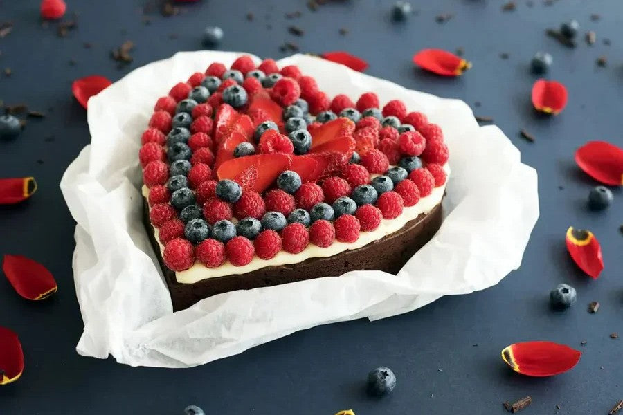 Herzförmiger Schokoladenkuchen mit Himbeeren, Blaubeeren und Erdbeeren, dekorativ auf weißem Papier arrangiert.