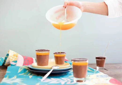 Schokoladen-Panna-Cotta mit Orangensoße, serviert in kleinen Gläsern auf buntem Tischtuch.