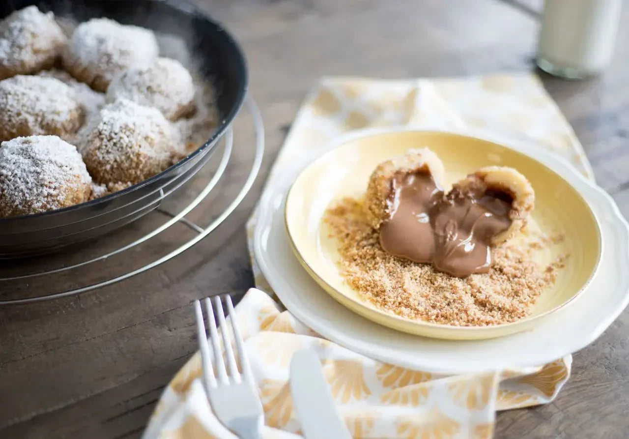 Geöffneter Nougatknödel auf Teller, gefüllt mit Schokolade und bestäubt mit Puderzucker, daneben weitere Knödel in Pfanne.