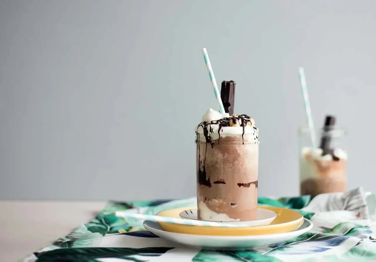Eisschokolade mit Haselnussstücken, Sahnehäubchen und Strohhalm in Glas, auf gelbem Teller mit tropischem Deko-Element.