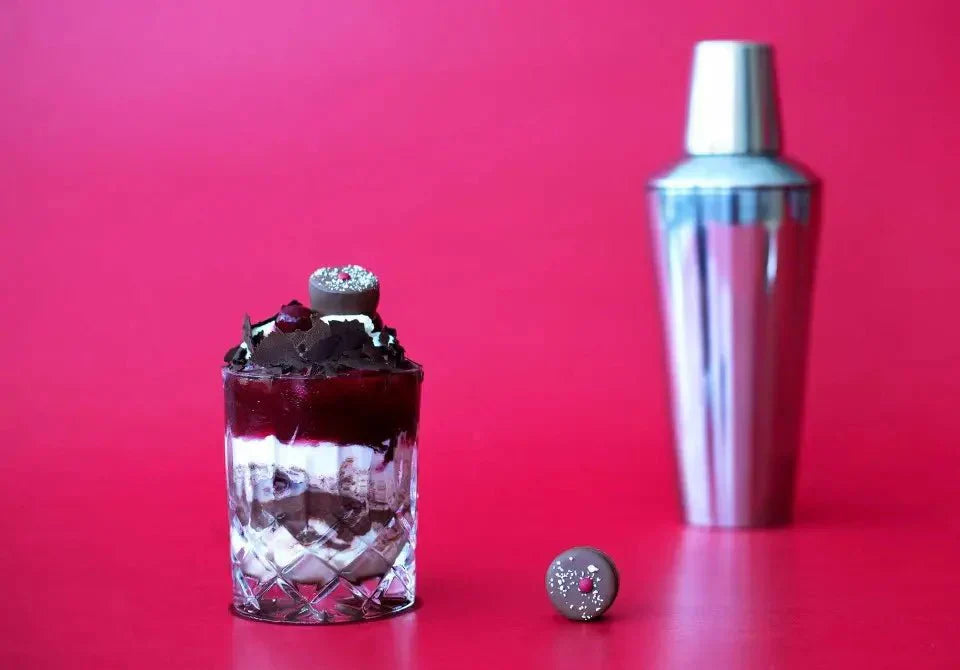 Schwarzwälder Kirschcocktail im Glas vor rotem Hintergrund, mit Schokoladendeko und Cocktail-Shaker.