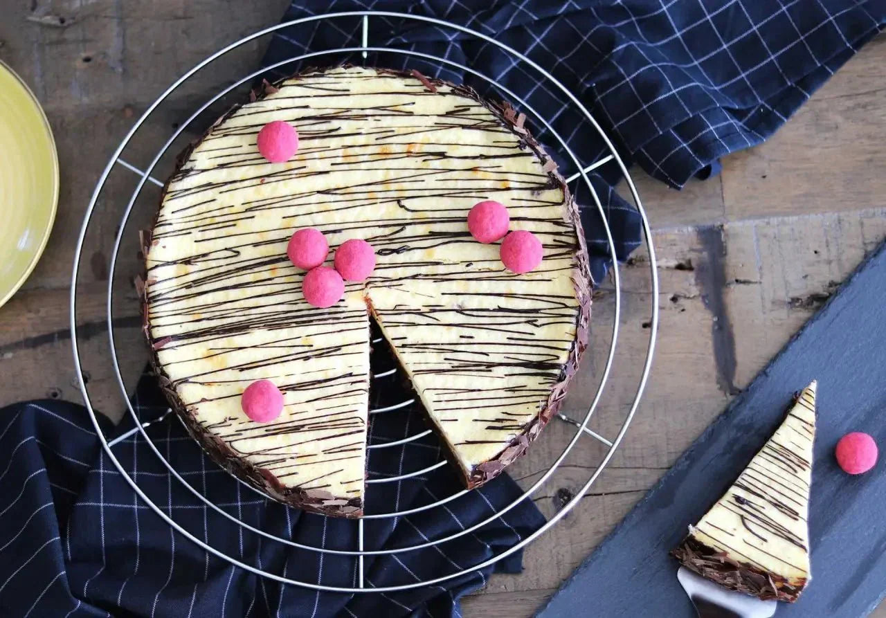 Käsekuchen mit Brownieboden, dekoriert mit Schokoladenstreifen und rosa Kugeln, auf einem Gitter mit Serviette.