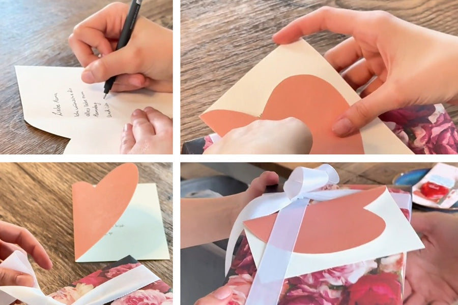 Person schreibt auf Herzkarte aus Papier und befestigt sie an Geschenk mit Blumenmuster und weißer Schleife.