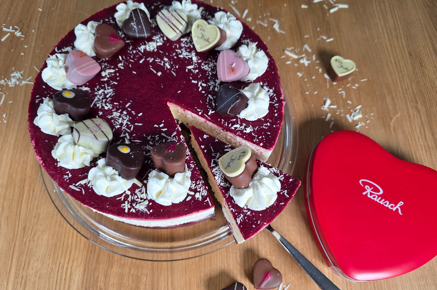 Kirschtorte mit Sahnetuff und Herzpralinen, daneben rote Herzschachtel von Rausch auf Holztisch.