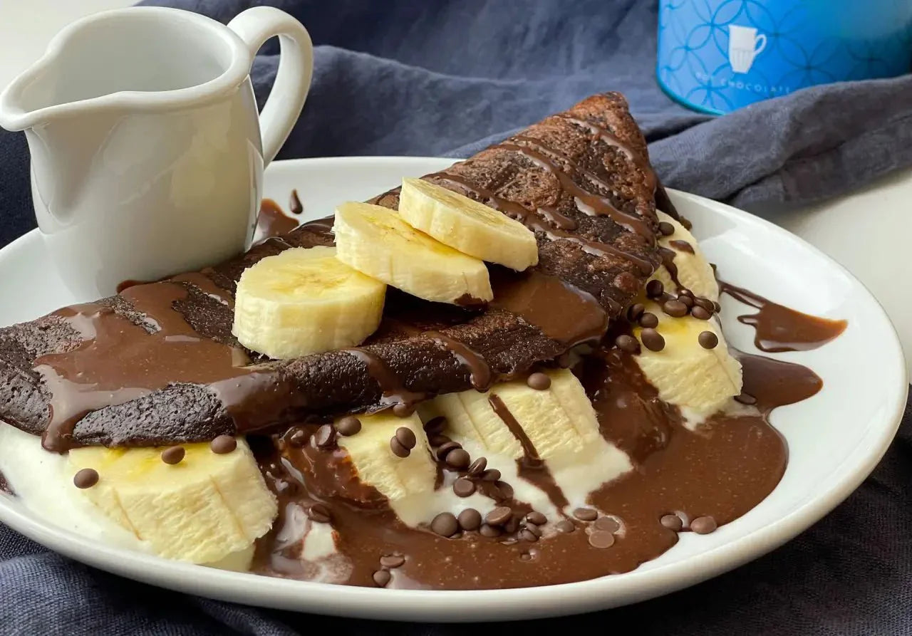 Schokopfannkuchen mit Bananenscheiben, Sahne und Kakaotropfen, serviert mit Kännchen auf weißem Teller.