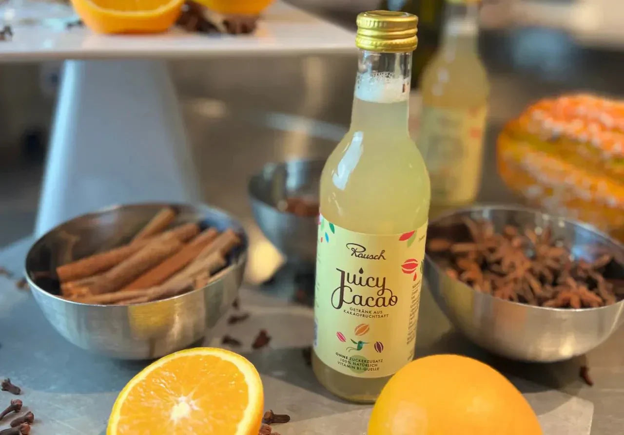 Flasche Juicy Cacao neben Orangenhälften, Gewürzen und Schalen auf einem Tisch.