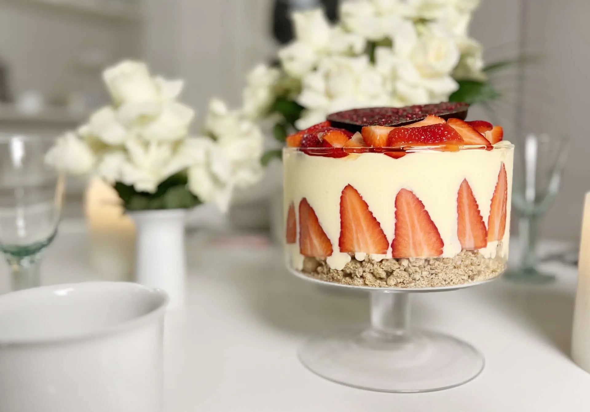 Elegantes Schichtdessert mit weißer Schokolade und Erdbeeren, dekoriert mit weißen Blüten im Hintergrund.