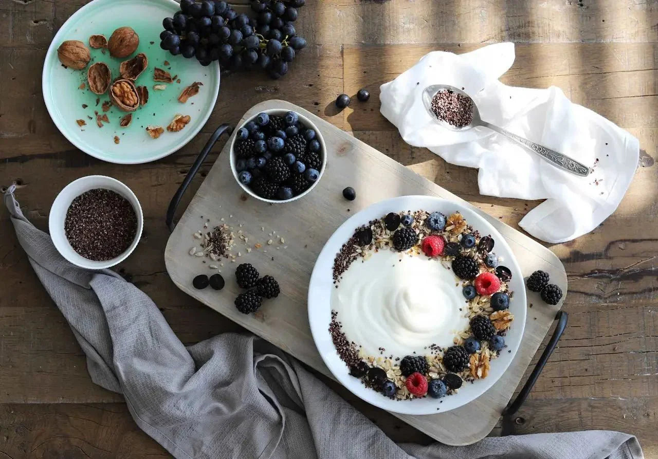 Schale mit Joghurt, Beeren und Kakaonibs auf Holzbrett, umgeben von Walnüssen und Trauben auf einem rustikalen Tisch.