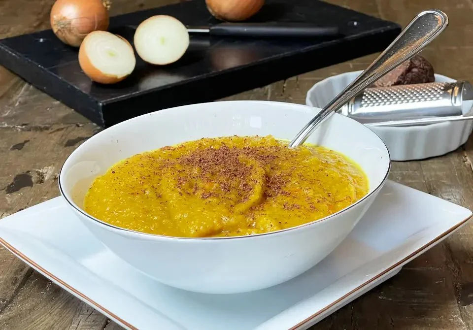 Schale mit gelber Kartoffelsuppe, bestreut mit Schokolade, auf Holztisch, Zwiebeln und Reibe im Hintergrund.