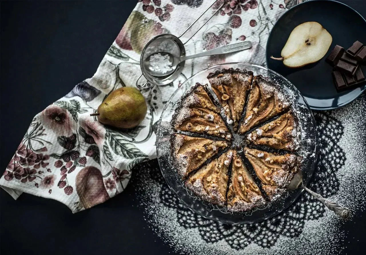 Schokoladen-Birnen-Tarte auf Spitzen-Unterlage, dekoriert mit Birne, Schokolade und Puderzucker auf dunkler Fläche.