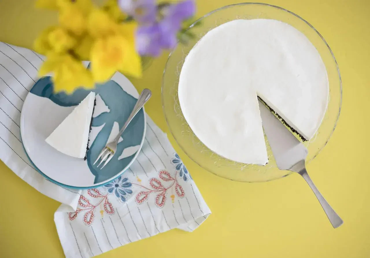 Weißer Triple Cheesecake mit Kuchengabel auf blumigem Teller neben Blumenstrauß auf gelbem Tisch.