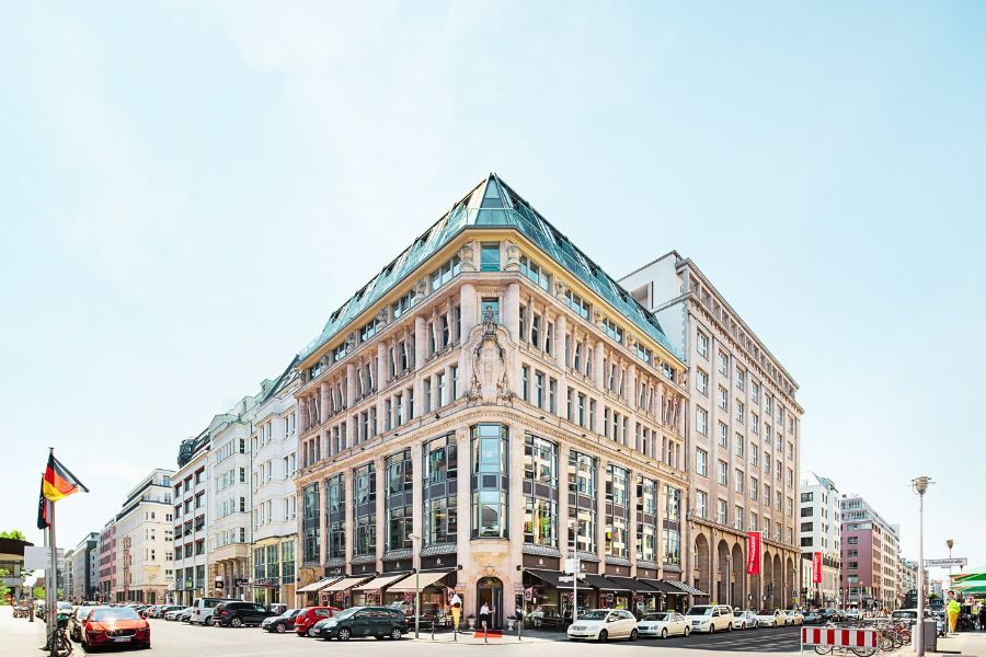 Historisches Berliner Gebäude in einer belebten Straßenszene mit Autos und Passanten, strahlend bei Tageslicht.