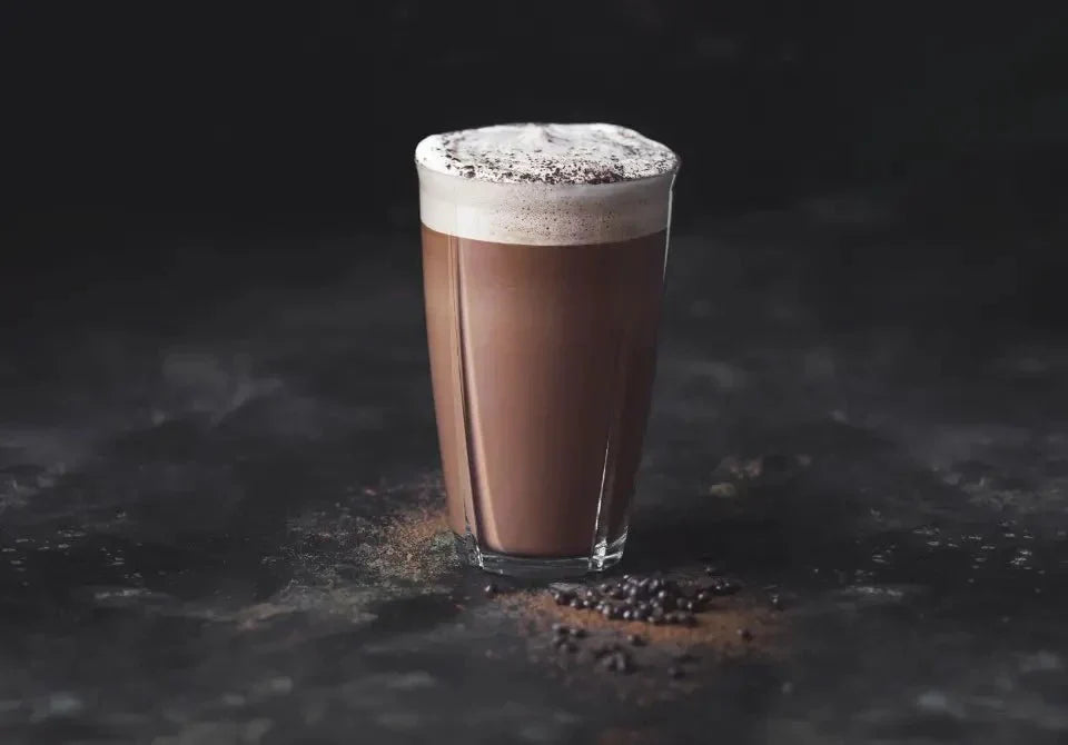 Ein Glas cremige Trinkschokolade mit Schaum und Schokoraspeln auf einer dunklen Oberfläche.