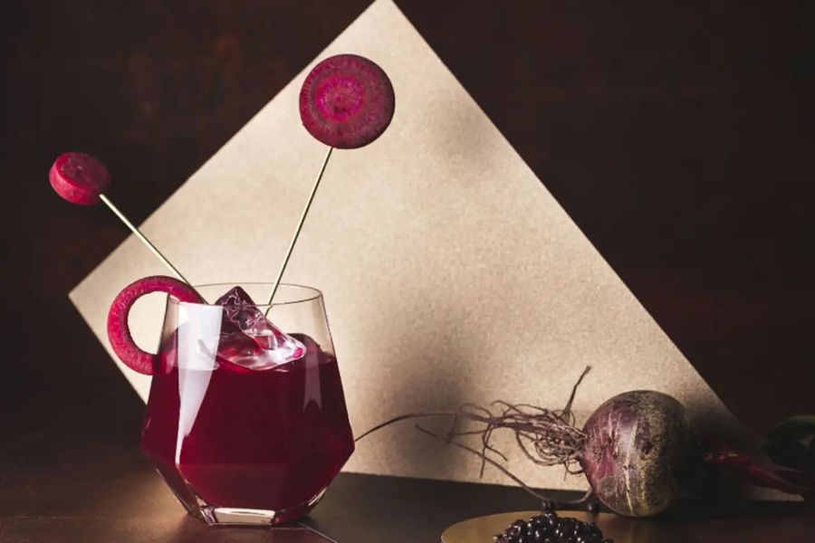 Rotes Getränk in modernem Glas, dekoriert mit Rote-Bete und Beeren, vor geometrischem Hintergrund.