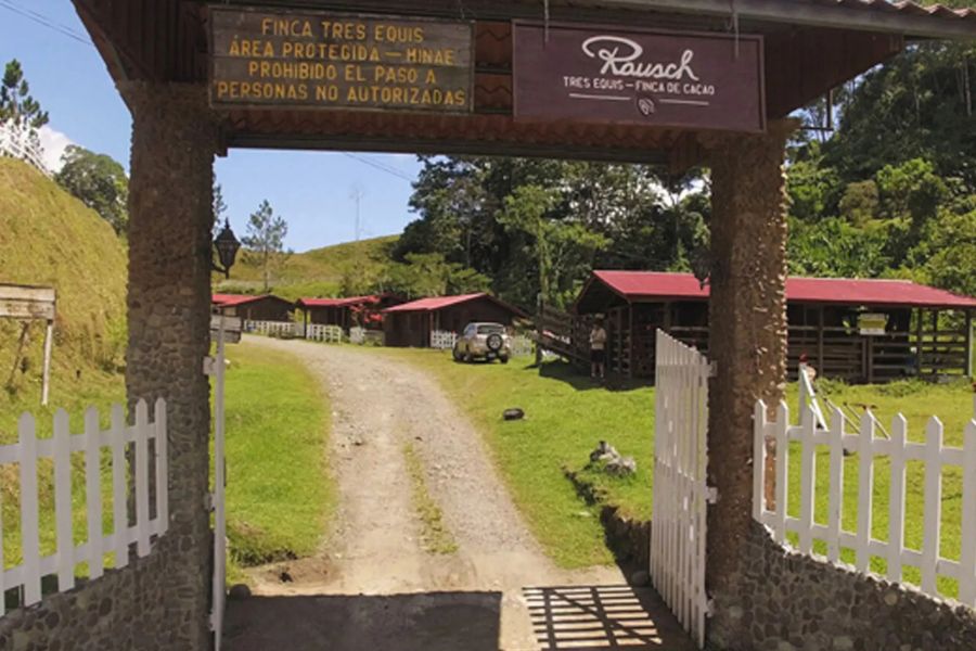 Eingang zur Finca Tres Equis von Rausch, umgeben von grünen Hügeln und Gebäuden mit roten Dächern.