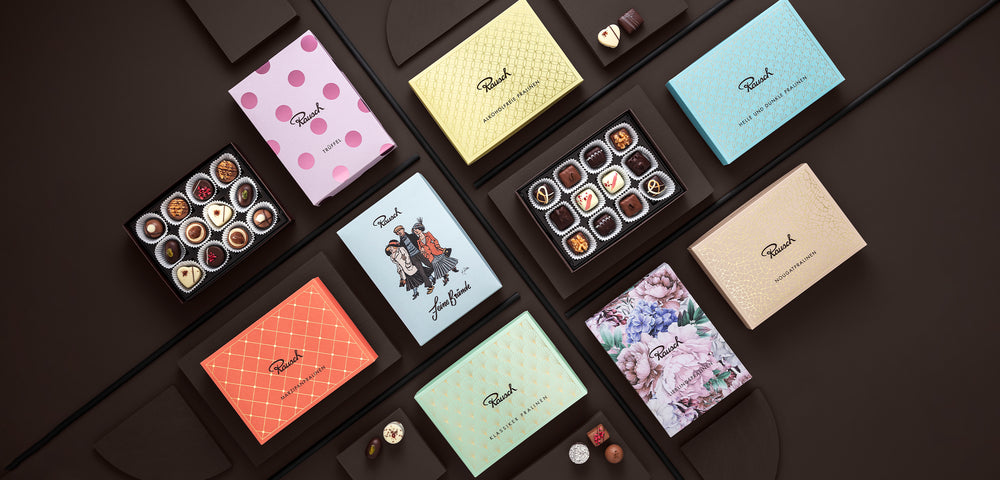 Verschiedene Pralinenschachteln von Rausch mit kunstvollen Designs und offenen Boxen mit Pralinen, auf dunklem Hintergrund.