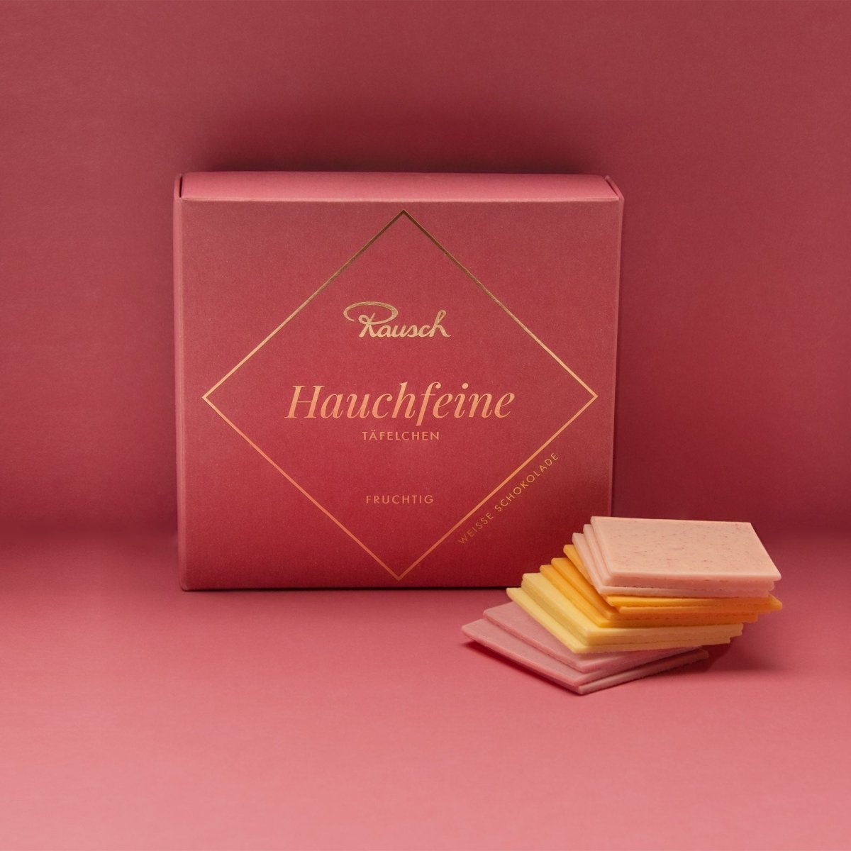 Rosane Schachtel Hauchfeine Täfelchen auf rosa Hintergrund mit 4 verschiedenfarbigen fruchtigen Täfelchen im Vordergrund.