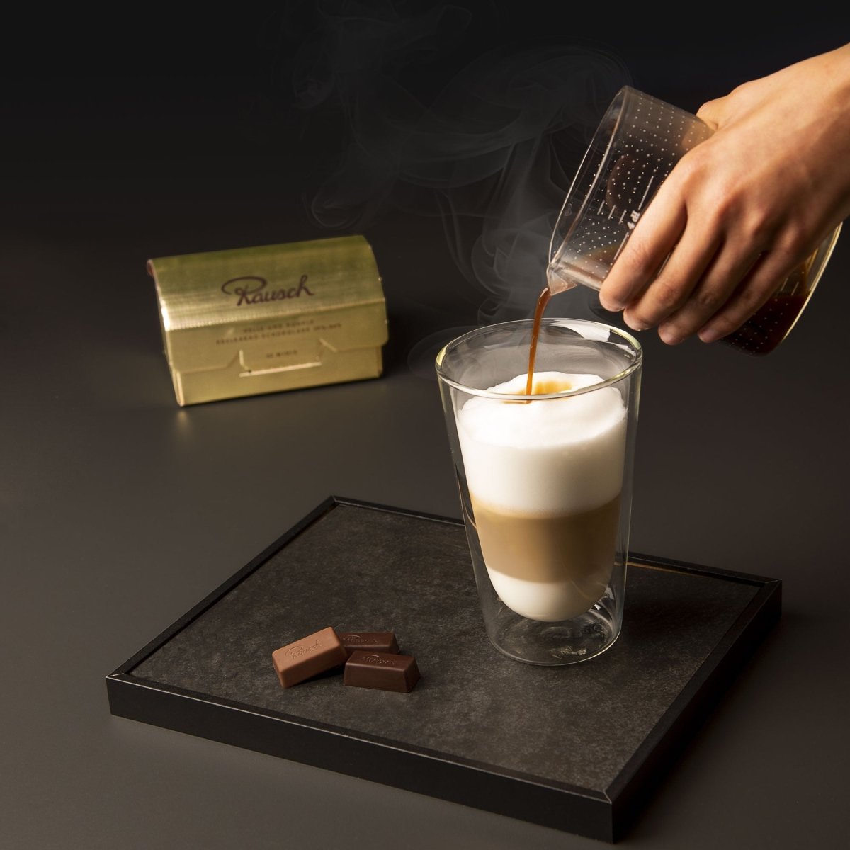 Eine Hand gießt Kaffee in einem Latte Macchiato, daneben liegen Rausch Plantagen Minis und im Hintergrund steht eine Goldbox.