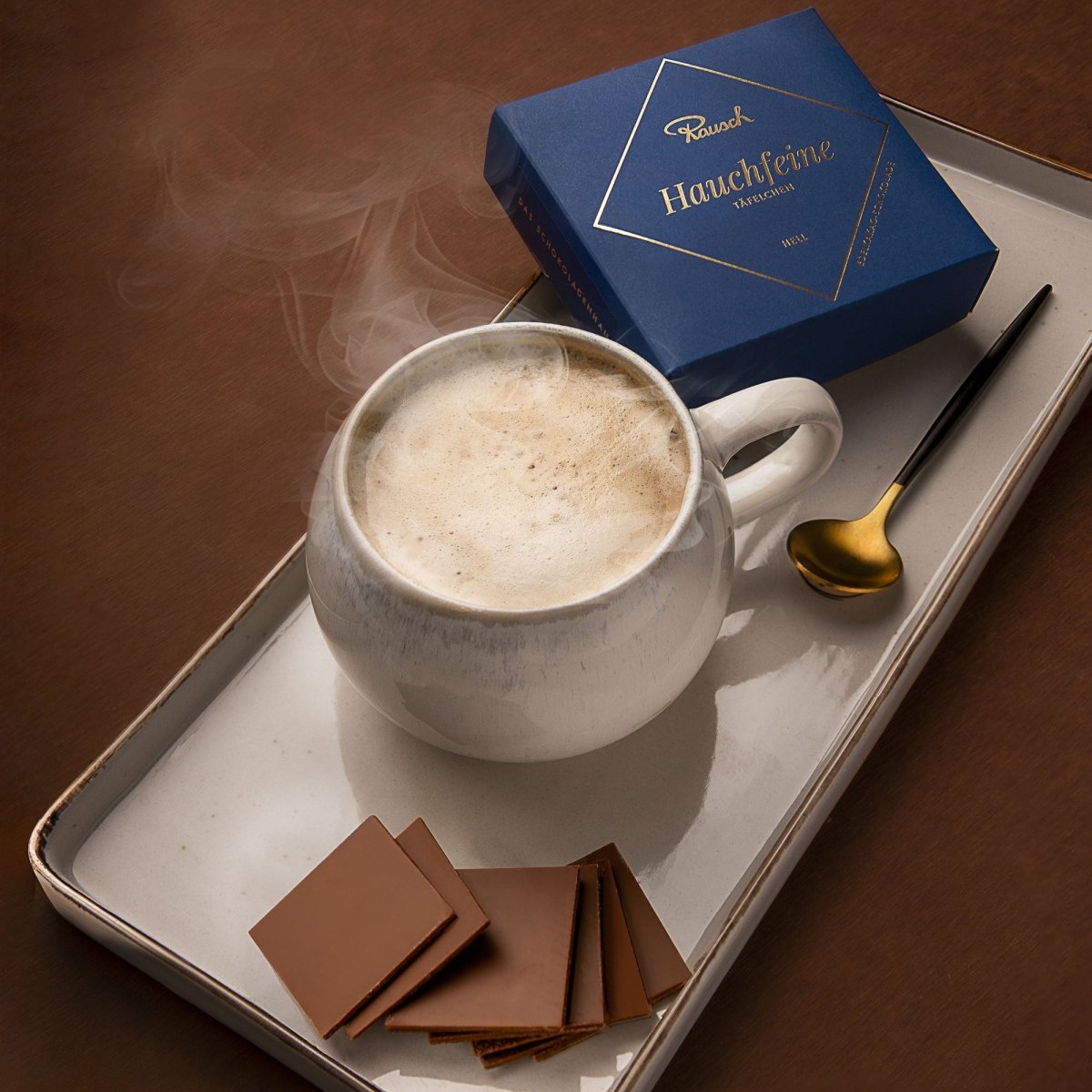 Eine Tasse Cappuccino und Hauchfeine Täfelchen in heller Edelkakao-Schokolade stehen auf einem rechteckigen Teller.