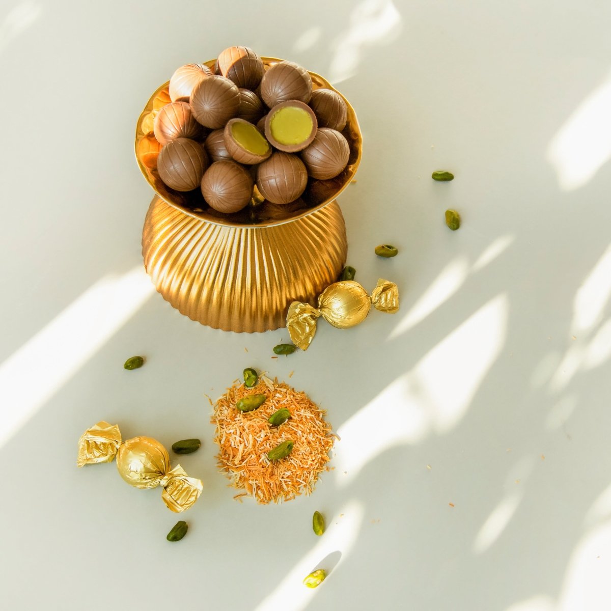 In einer goldenen Schüssel liegen viele Dubai-Pralinenkugeln. Davor liegen zwei gold verpackte Kugeln und einzelne Pistazien auf Kadayif.